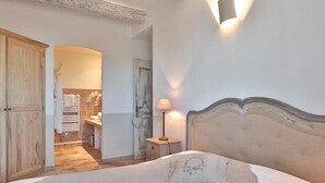 Quarto com uma cama grande, mesa de cabeceira com abajur, guarda-roupa de madeira e banheiro visível por uma porta aberta.
