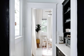A narrow hallway with white walls and a door leading to a dining area. A potted plant and a white chair are visible in the dining area.