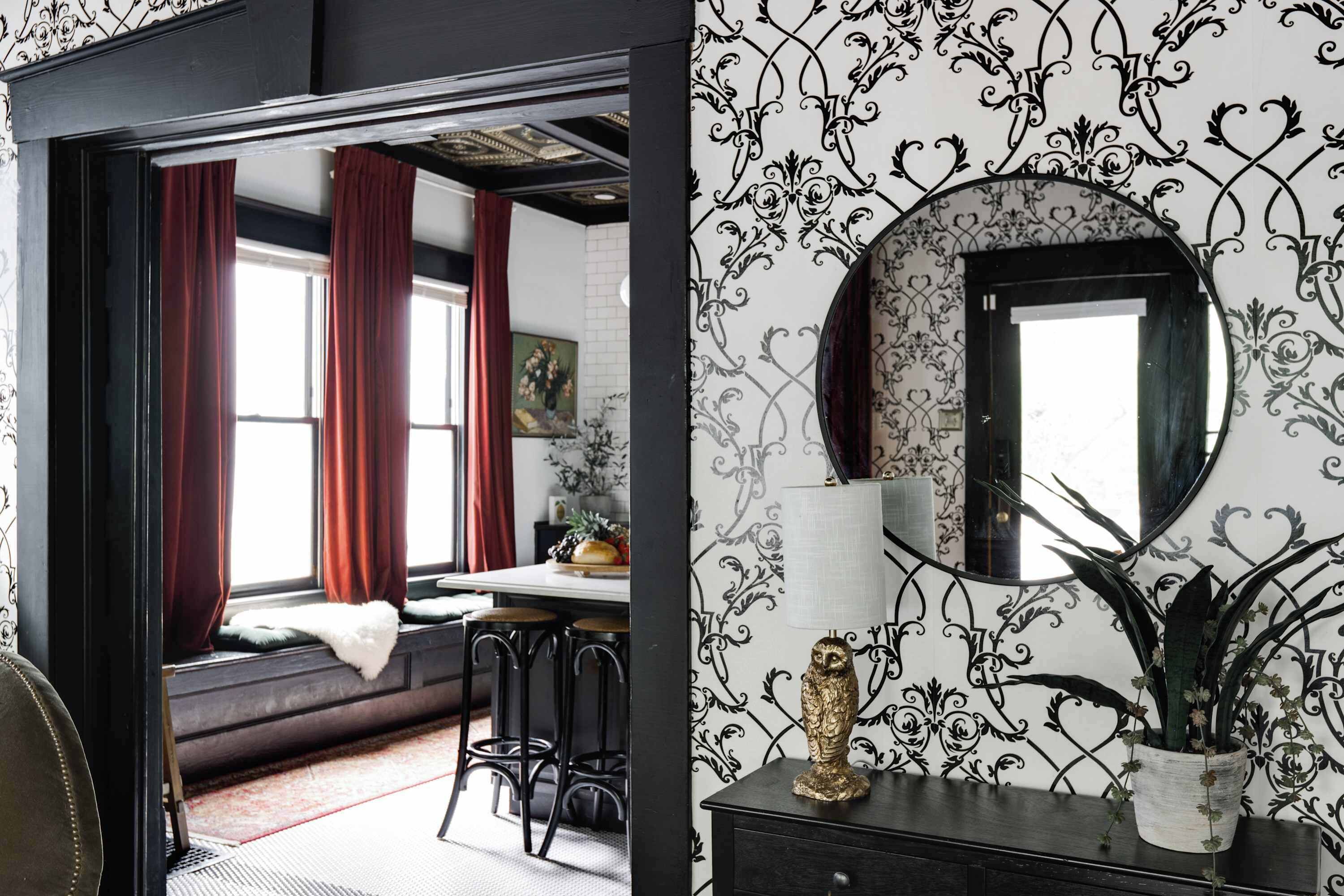 A room with a black and white patterned wallpaper, a round mirror, a potted plant, and a white lamp on a dark wooden dresser.