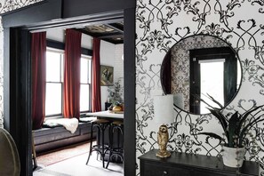 A room with a black and white patterned wallpaper, a round mirror, a potted plant, and a white lamp on a dark wooden dresser.