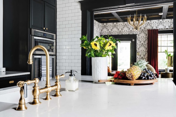 Eine moderne Küche mit schwarzen Schränken, einer weißen Fliesenfassade, einem Edelstahlbackofen, einem goldenen Wasserhahn und einem Strauß Sonnenblumen auf der Arbeitsplatte.