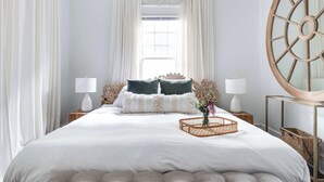 Une chambre à coucher bien aménagée, avec un grand lit, deux tables de chevet avec des lampes, un miroir décoratif et une fenêtre avec des rideaux.