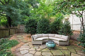A curved wicker sofa with a round table in a garden area with stone paving, surrounded by dense foliage and a wooden fence.