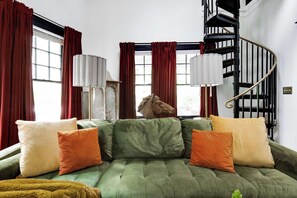 A green sofa with orange and white cushions, a horse head sculpture, and a black spiral staircase.
