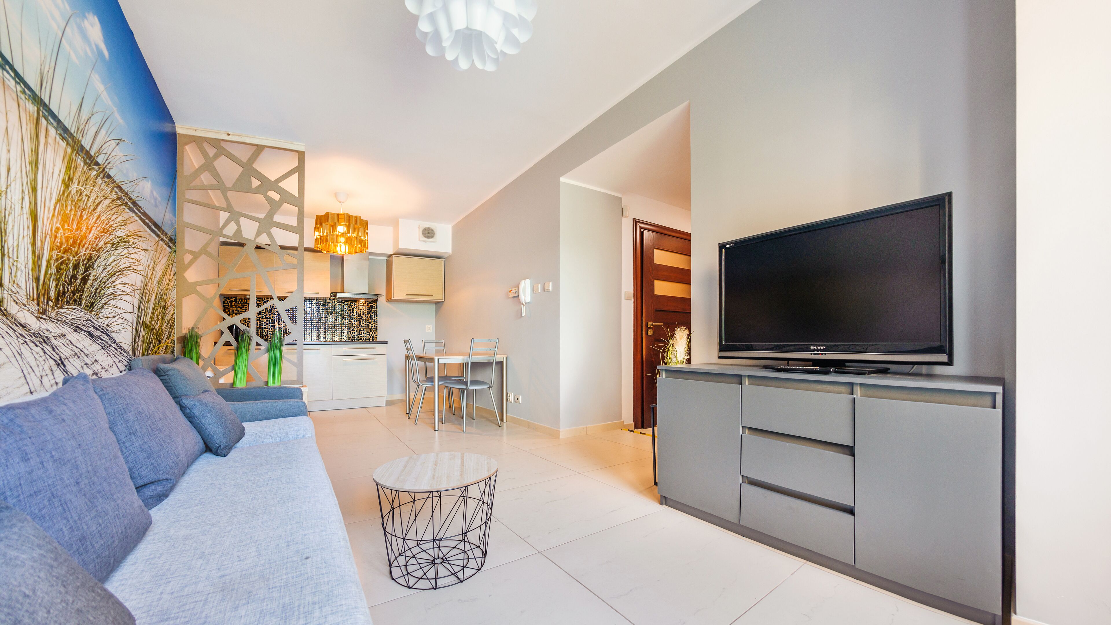 A modern living room with a grey sofa, a TV on a grey cabinet, and a kitchen in the background.