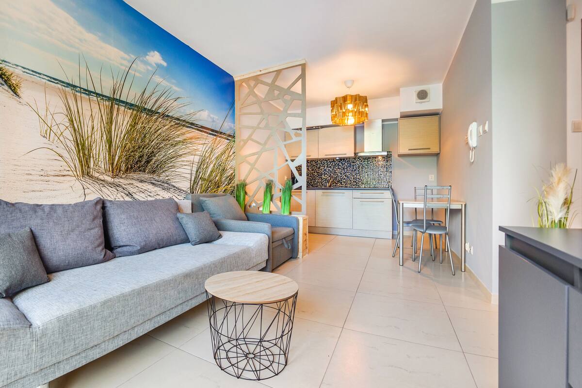 Ein modernes Wohnzimmer mit einem grauen Sofa, einem runden Holznachttisch und einer Küchenzeile mit weißer Arbeitsplatte und einer Pendelleuchte.