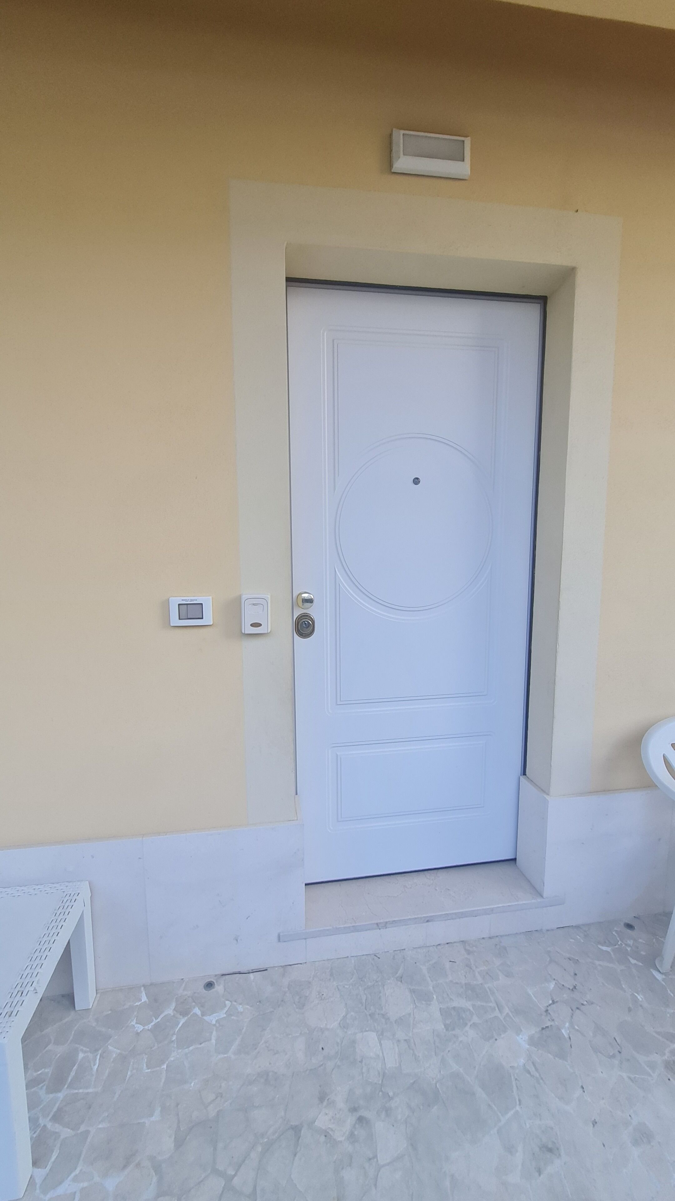 Une porte blanche ornée d’un motif circulaire, un petit luminaire au-dessus, et un thermostat fixé au mur sur la gauche.