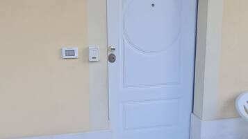 Une porte blanche ornée d’un motif circulaire, un petit luminaire au-dessus, et un thermostat fixé au mur sur la gauche.