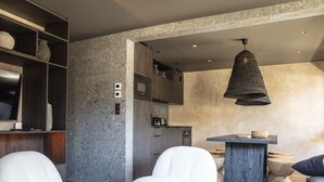 A modern living room with a round coffee table, two white armchairs, a bookshelf, and a kitchen area with a hanging light fixture. - Apartment FERRONNE by JAPANDI home (Megève)