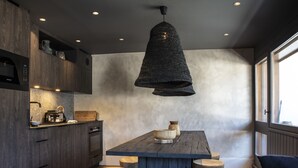 A modern kitchen with dark wood cabinets, a central island with stools, a hanging light fixture, and a view through a window. - Apartment FERRONNE by JAPANDI home (Megève)