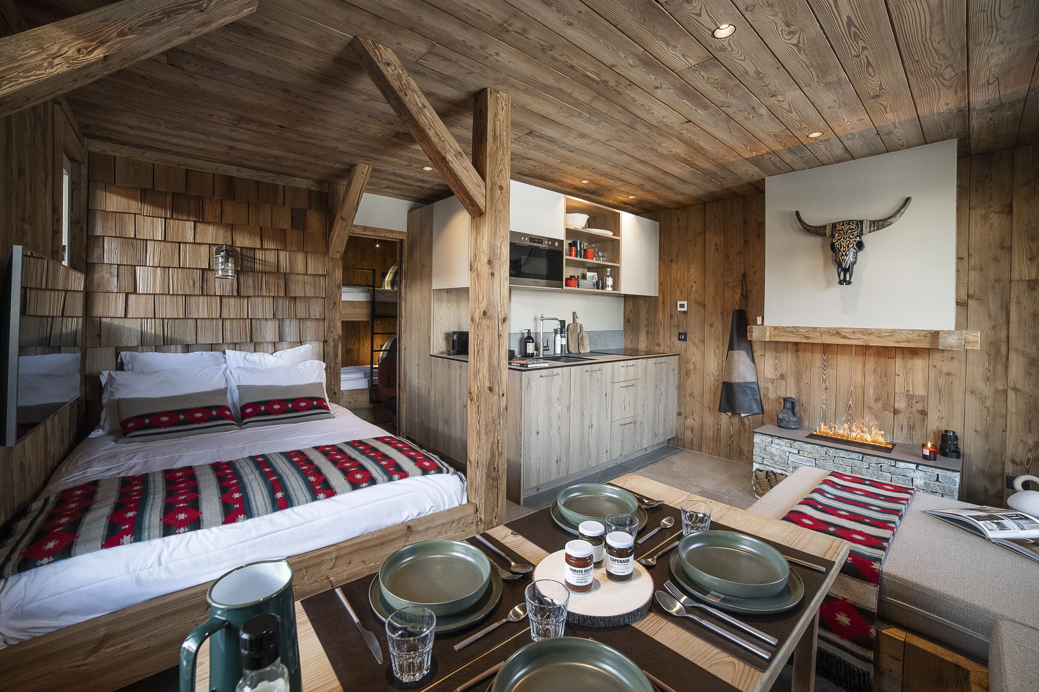 Un interior de madera con una cama, una mesa de comedor para dos, una zona de cocina y una decoración de cabeza de toro montada en la pared.