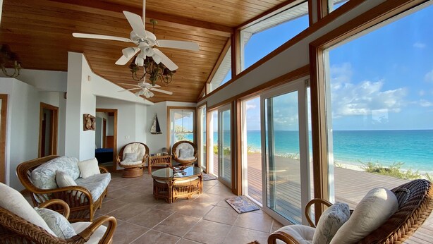 A spacious room with wicker furniture, a ceiling fan, and large windows overlooking a beach.