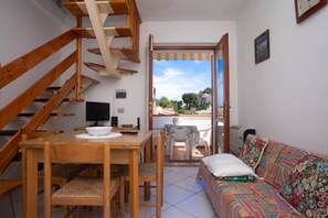 A room with a wooden staircase, a dining table with chairs, a sofa, and a view through an open door.