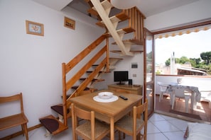 A room with a wooden staircase, a dining table with chairs, a television, and a balcony with outdoor seating.