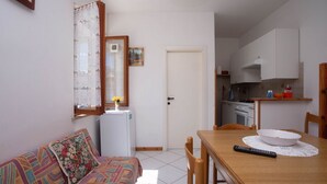 A small, well-lit room with a sofa, a dining table with chairs, a refrigerator, a kitchen area, and a door. - M291 - Marcelli, apartment with terrace and reserved parking (Marcelli)