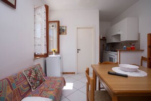 A small, well-lit room with a sofa, a dining table with chairs, a refrigerator, a kitchen area, and a door.