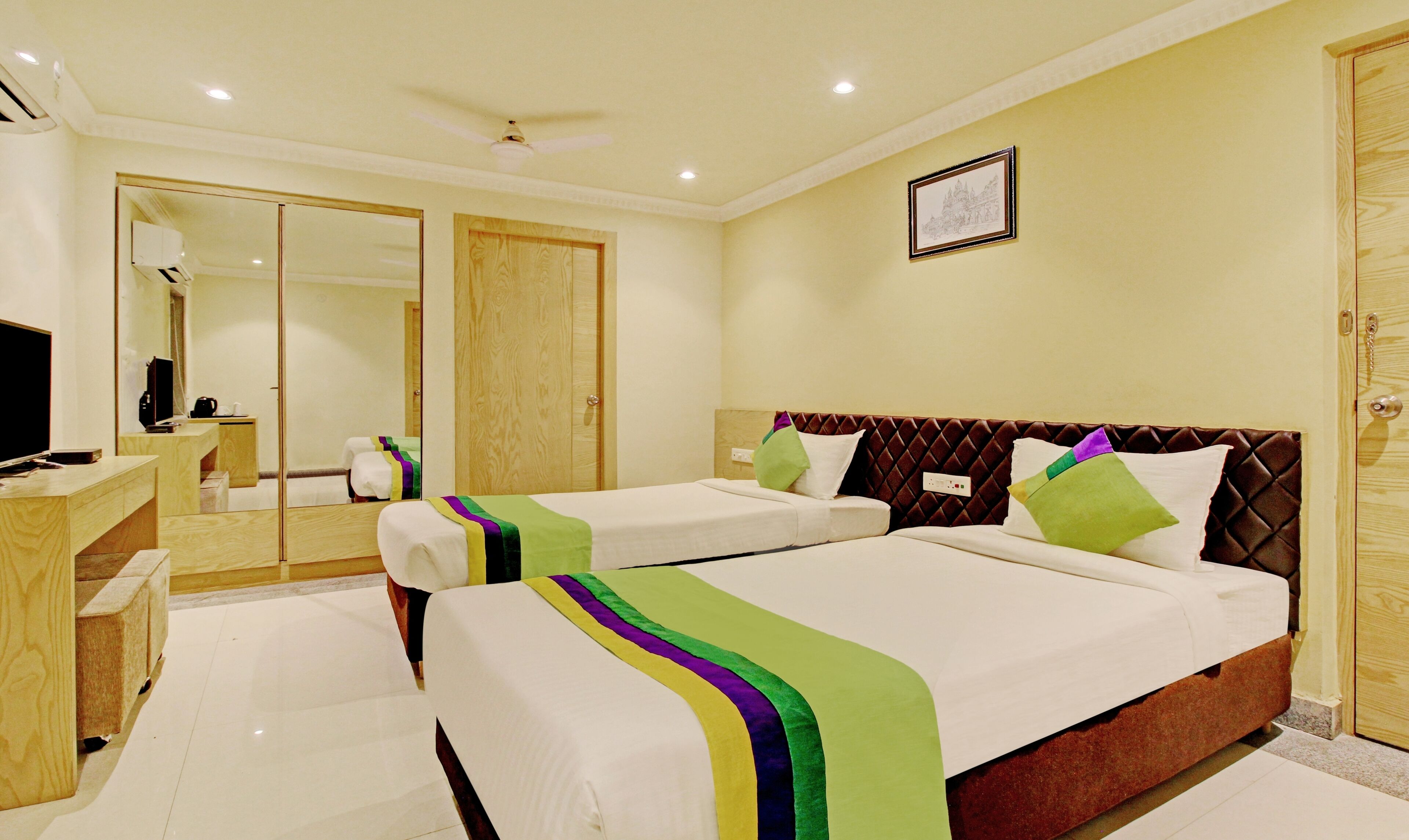 A hotel room with two beds, a desk, a mirror, and a framed picture on the wall.
