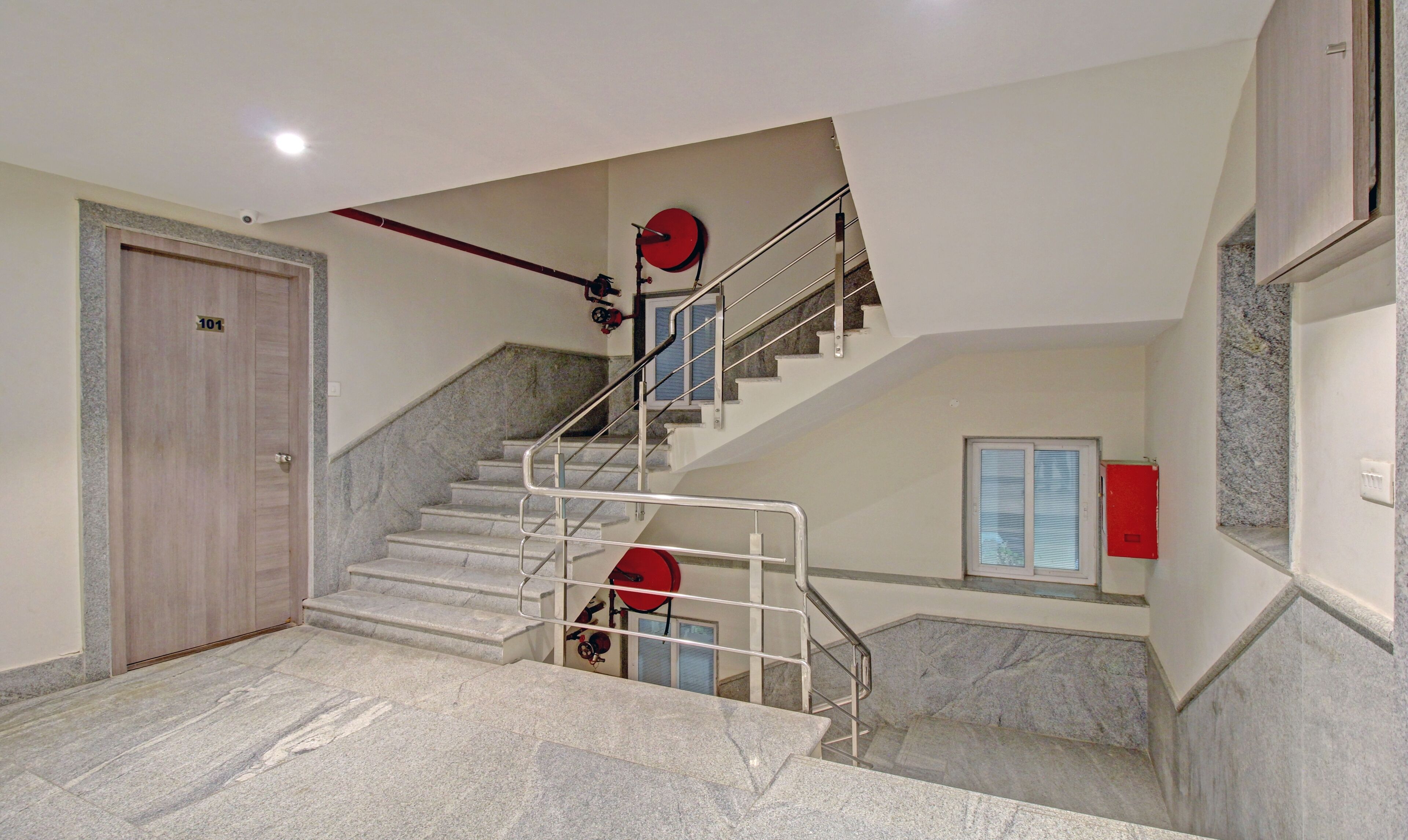 Una escalera con peldaños de mármol y barandillas de metal, una puerta de madera con una placa de número, y un letrero de salida de emergencia en rojo.