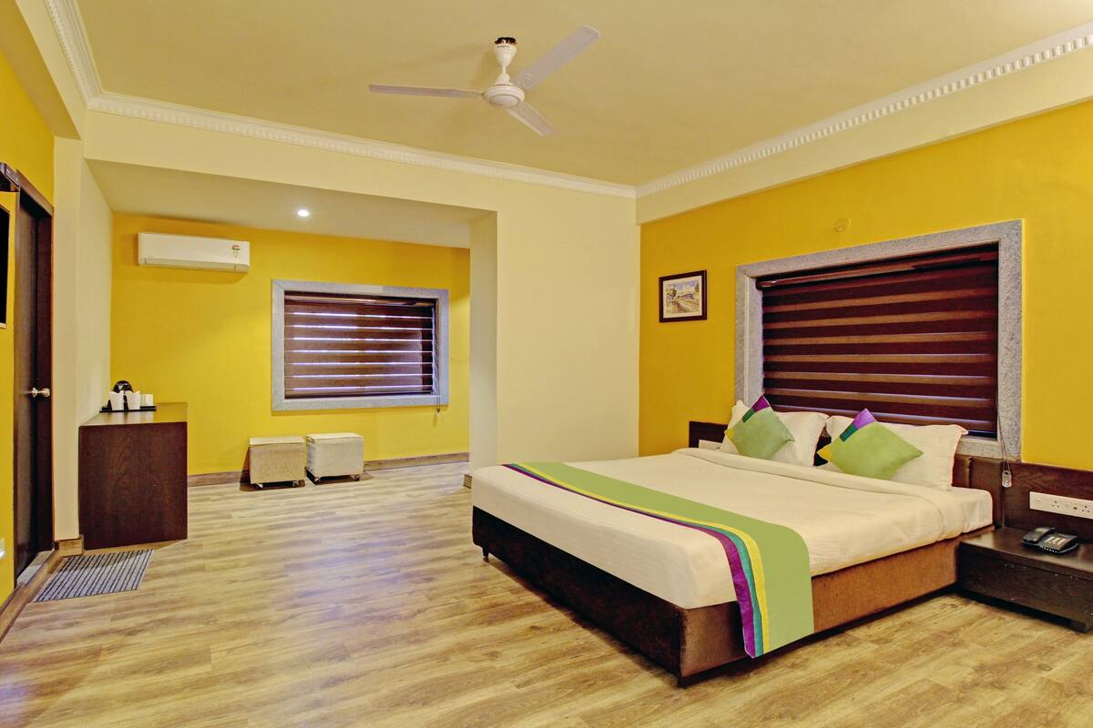 A hotel room with a bed, a desk, a chair, a window with blinds, a ceiling fan, and a wall-mounted air conditioner.