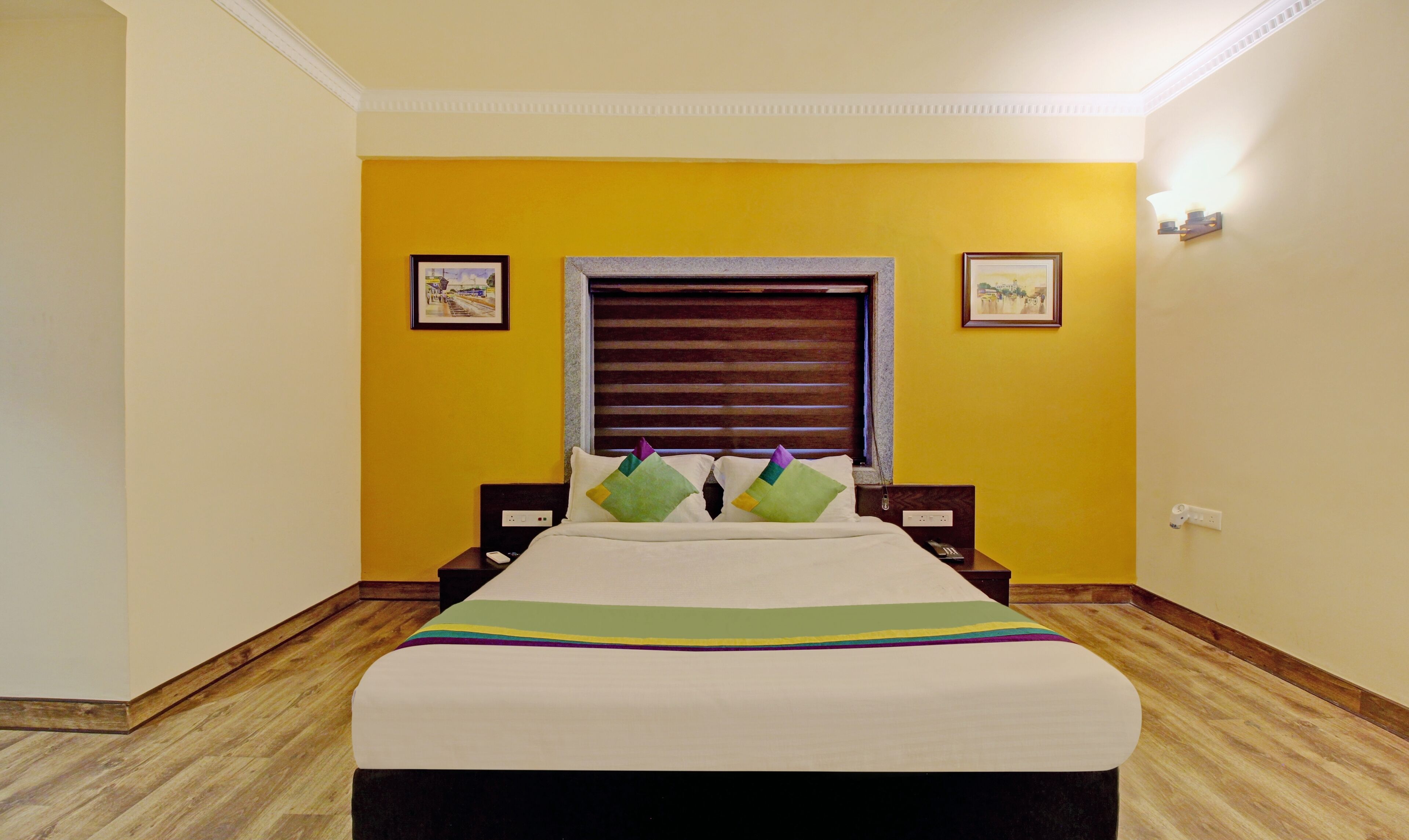Quarto com uma cama, janela com persianas, duas fotos emolduradas nas paredes e piso de madeira.