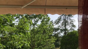 A covered outdoor area with a table and chairs, a decorative statue, and a view of a garden with trees and a pathway. - Maison D'artiste Atypique (Saint-Hippolyte-du-Fort)