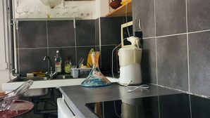 A kitchen with a black tiled backsplash, a white countertop, a sink, and a stove. There is a shelf with glassware and a painting on the wall. - Maison D'artiste Atypique (Saint-Hippolyte-du-Fort)