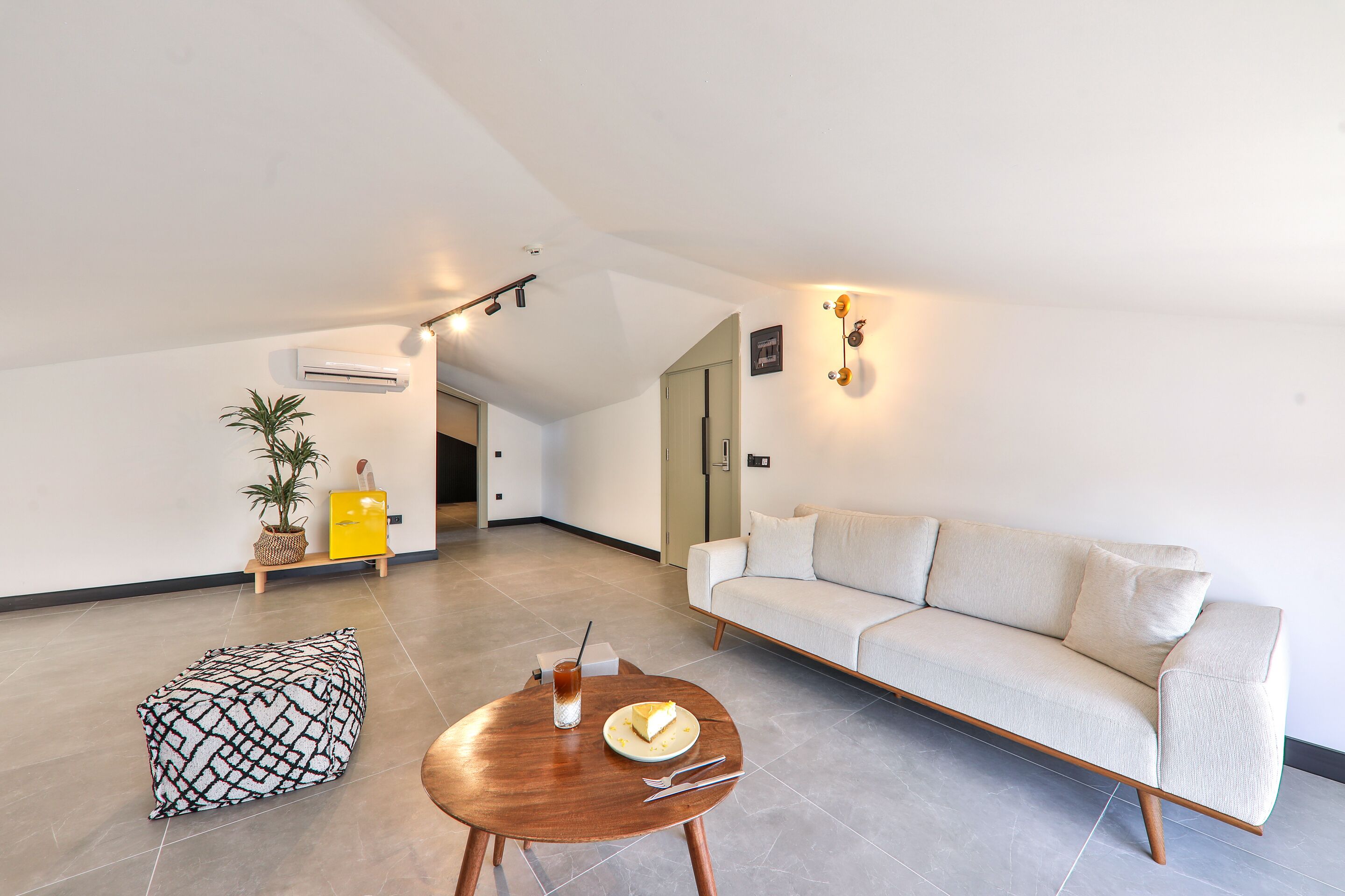 A modern living room with a sofa, a wooden coffee table with a plate and utensils, a patterned ottoman, and a potted plant.