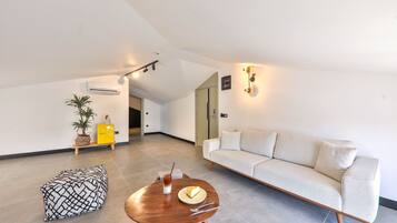 A modern living room with a sofa, a wooden coffee table with a plate and utensils, a patterned ottoman, and a potted plant.