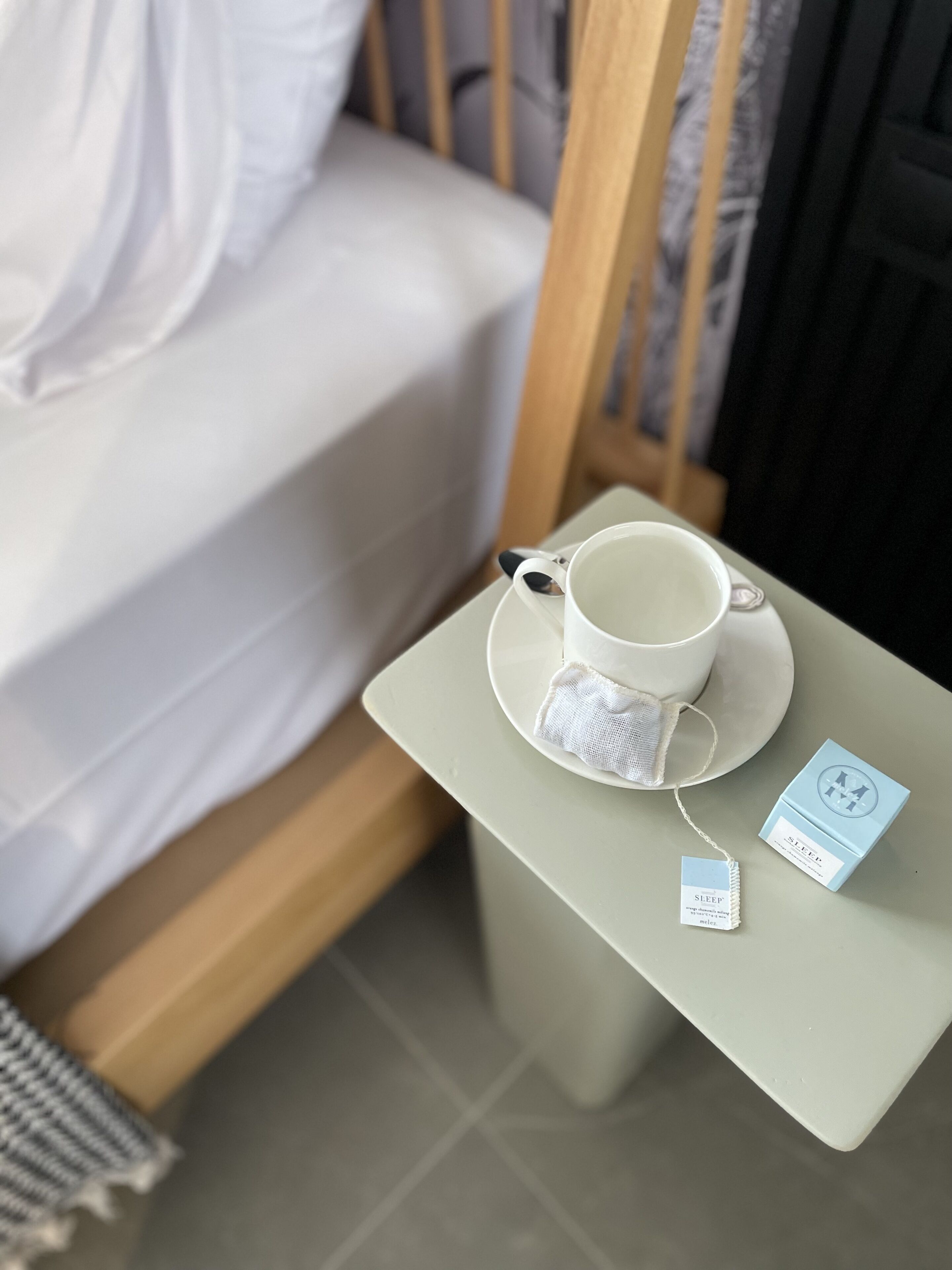 Una cama bien hecha con ropa blanca, una taza y platillo blancos sobre una mesita, y una bolsita de té y una caja etiquetada “SLEEP” junto a la taza.