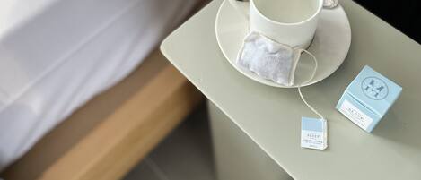 Una cama bien hecha con ropa blanca, una taza y platillo blancos sobre una mesita, y una bolsita de té y una caja etiquetada “SLEEP” junto a la taza.