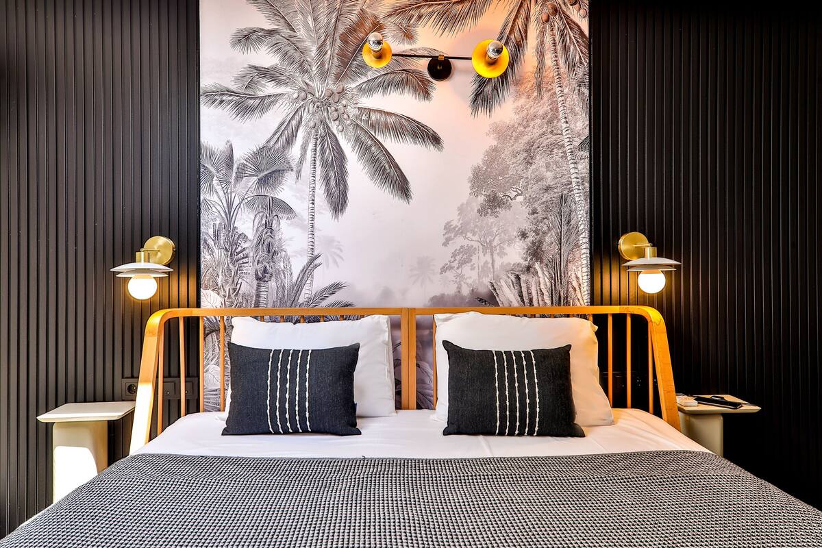 Una habitación de hotel con cama, dos mesitas de noche, un mural en la pared con palmeras y dos lámparas de pared.
