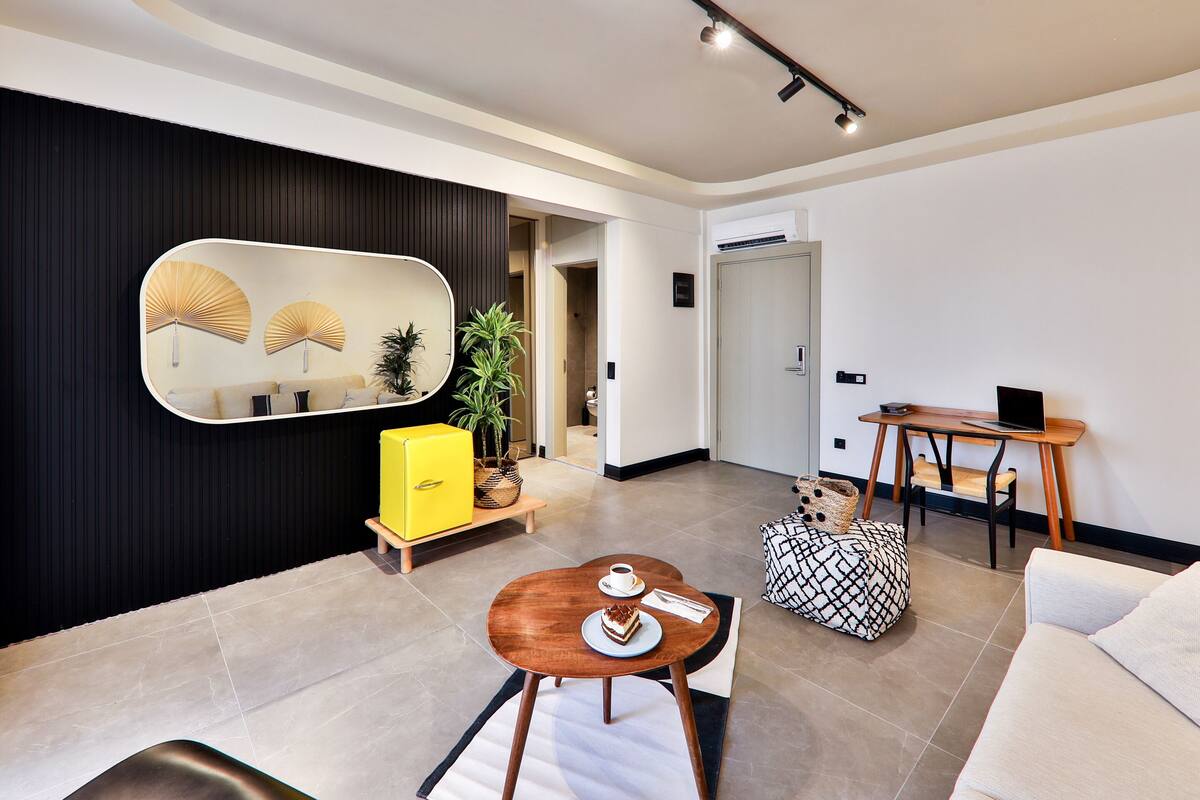Una sala de estar moderna con un sofá, una mesa de centro, un aparador amarillo, un espejo y una pequeña mesa de madera con una computadora portátil.