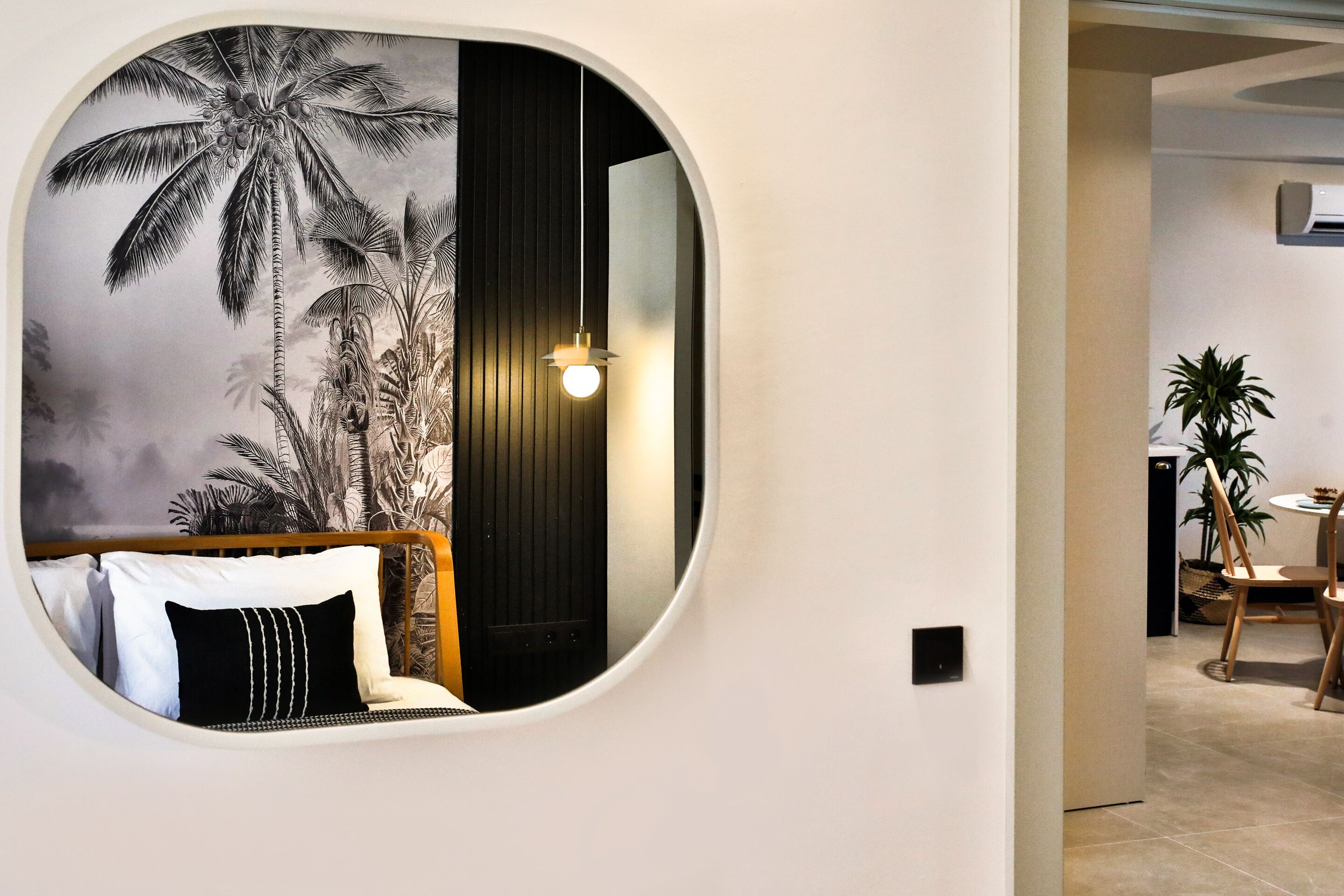 A circular mirror reflecting a bed with white and black pillows, a wall with a palm tree mural, and a hanging light fixture.