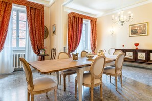 Una sala da pranzo con un lungo tavolo di legno, sei sedie, tende rosse decorate con motivi, un lampadario e un camino con soprammobili.