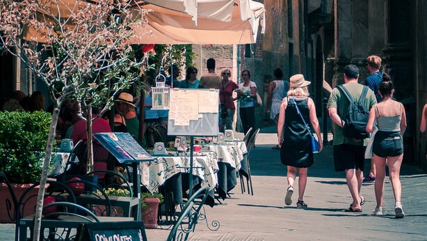 Een smalle straat met terrasjes, een naambord en een grote boog.