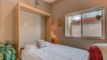 Un dormitorio con una cama, una ventana que da a una casa, un marco de madera, una planta y una silla roja.