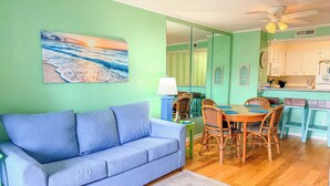 A living room with a blue sofa, dining table with chairs, a kitchen area, and a ceiling fan. - Stay at Tilghman Beach & Racquet Club 241 a second-row 3BR condo just steps from the sand with two balconies, pools, and tennis courts. (North Myrtle Beach)