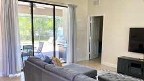 Una sala de estar con un sofá seccional gris, un televisor de pantalla plana y un puerta corredera de vidrio que da a un patio exterior con una parrilla.