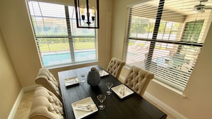 Comedor con mesa de madera oscura, sillas tapizadas en tono beige y un jarrón como centro de mesa. Se observan dos ventanas con persianas, y un ventilador de techo.