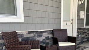 Un patio con dos sillas de mimbre, una mesa pequeña, un muro de piedra y una puerta con un letrero de número.