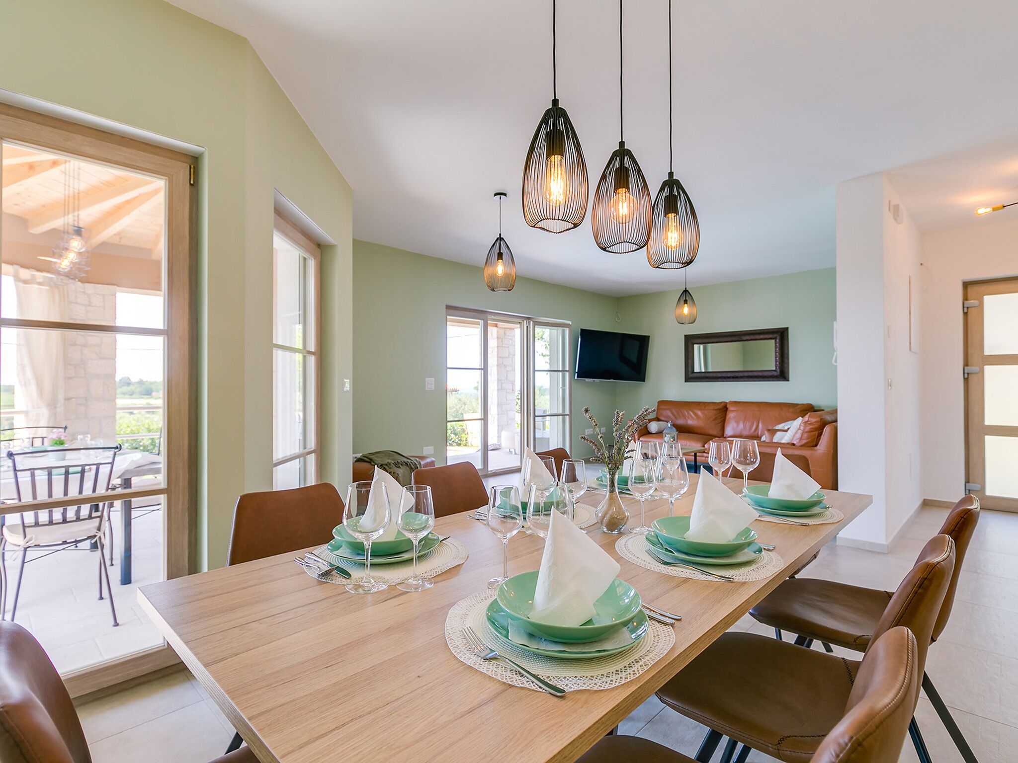 Un coin repas avec une longue table en bois dressée pour le repas, un canapé en cuir marron et un téléviseur fixé au mur.