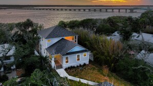 A two-story house with a blue roof, surrounded by dense greenery and a view of a bridge over a body of water during sunset. - Gorgeous Waterfront w/ Golf Cart! 4 bds, 3.5 ba! Sleeps 12 (St. Augustine)
