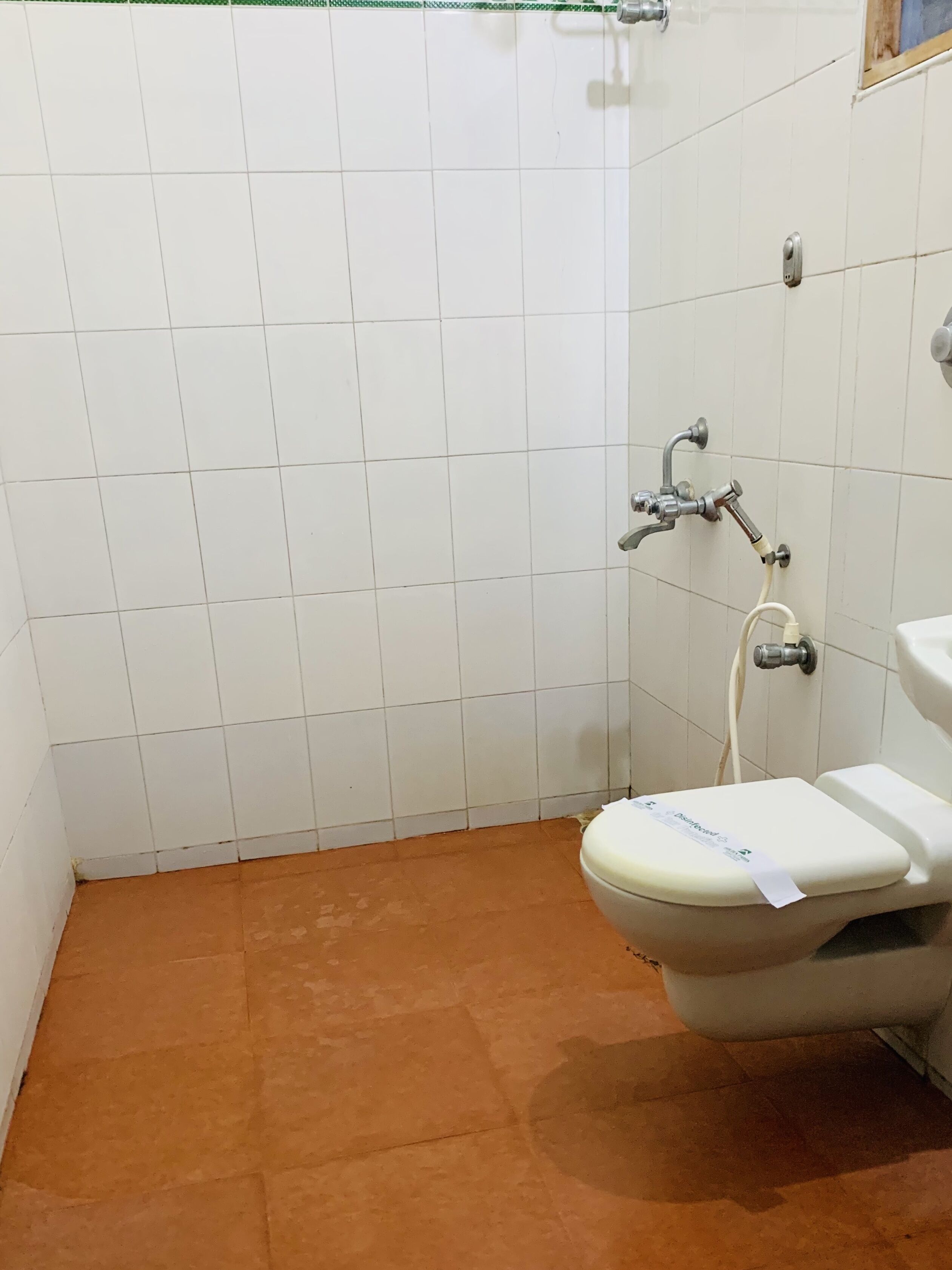 Une petite salle de bain compacte avec des murs carrelés de blanc et un sol marron. Elle comprend des toilettes et une douche avec un robinet.