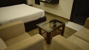 Une chambre d’hôtel comprenant un lit, deux canapés, une table basse en verre et une petite étagère avec une télécommande.
