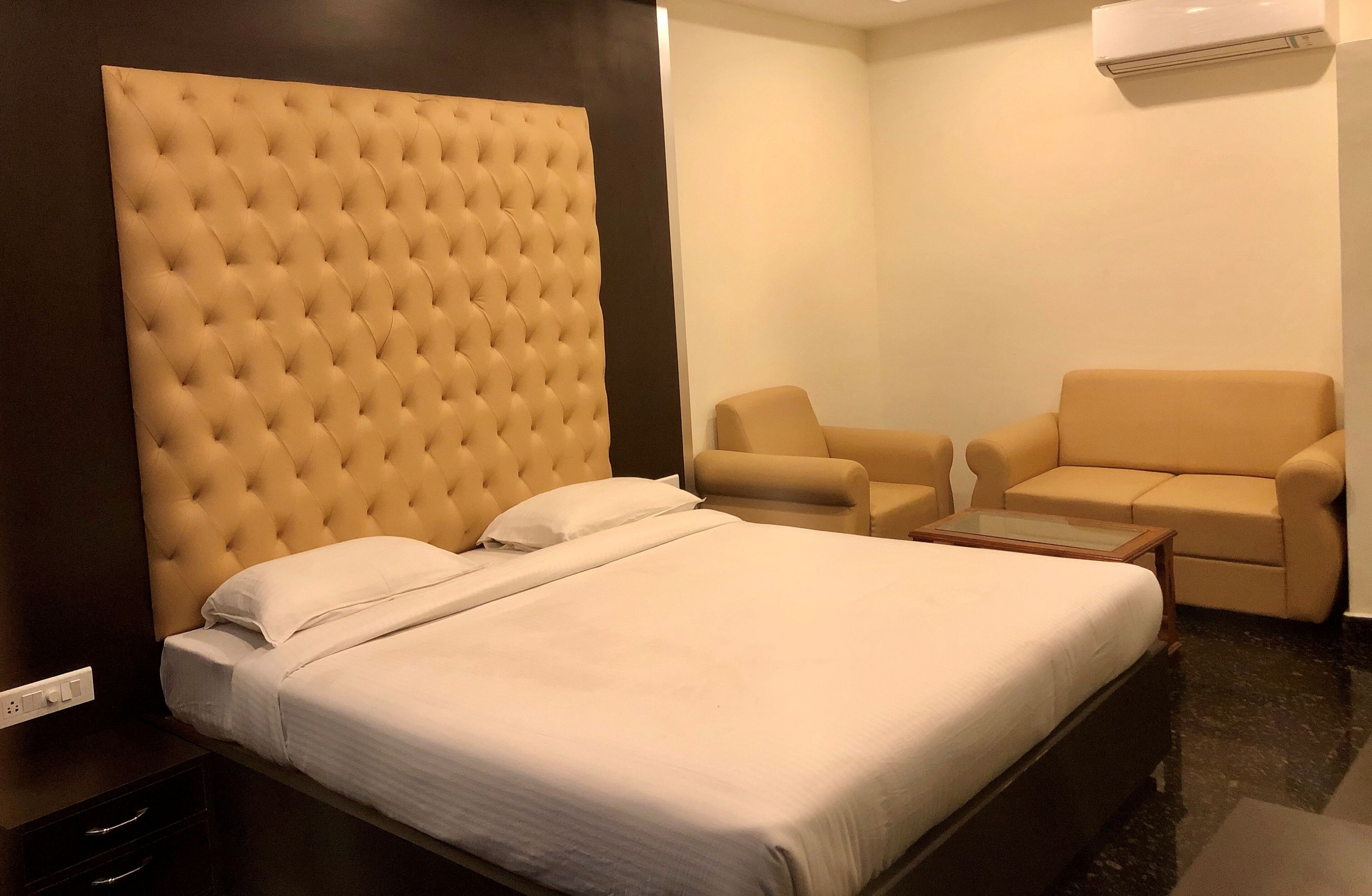 Ein Schlafzimmer mit einem großen, gepolsterten Kopfteil, einem Bett mit weißen Bettwäsche, zwei beigen Sesseln und einem kleinen Tisch.