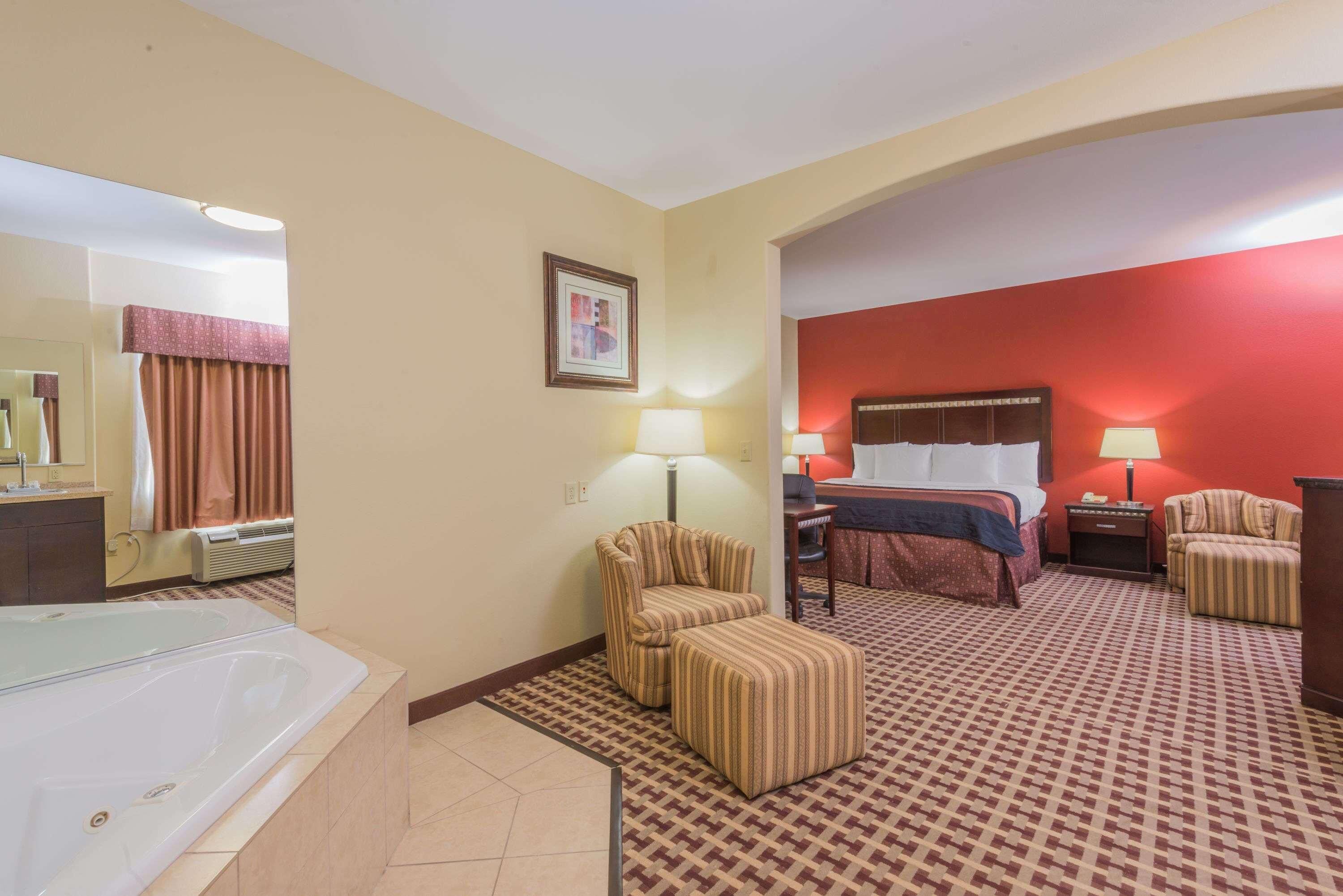 A hotel room with a large bed, a sitting area with a chair, a small table, a lamp, a mirror, and a bathtub.