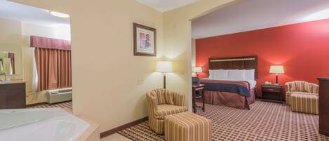 A hotel room with a large bed, a sitting area with a chair, a small table, a lamp, a mirror, and a bathtub.