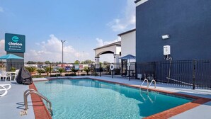A swimming pool with a blue and red border, a sign for Clarion Pointe by Choice Hotels, and a fenced patio area with outdoor seating. - Clarion Pointe Sulphur - Lake Charles (Sulphur)