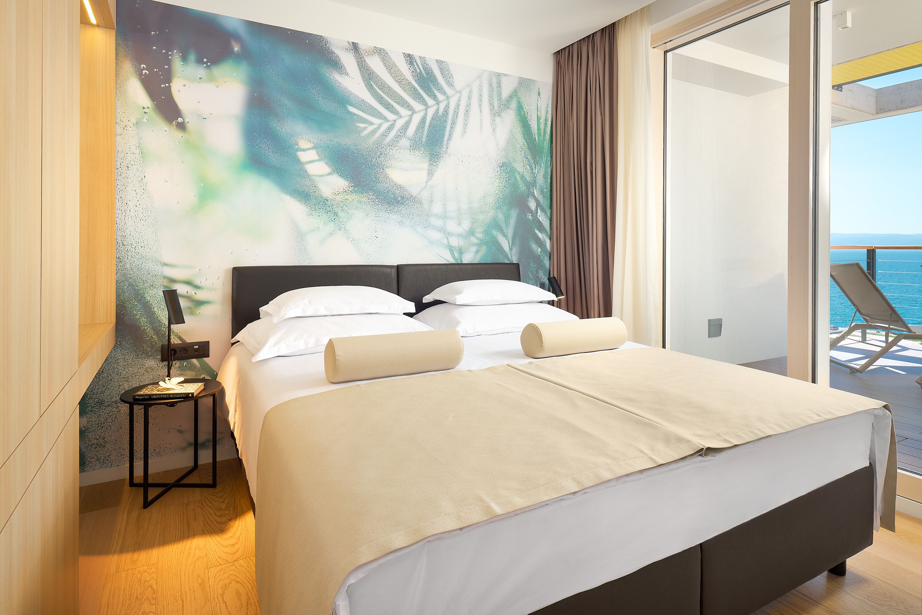 A modern hotel room with a large bed, a small side table, and a view of the sea through a balcony.