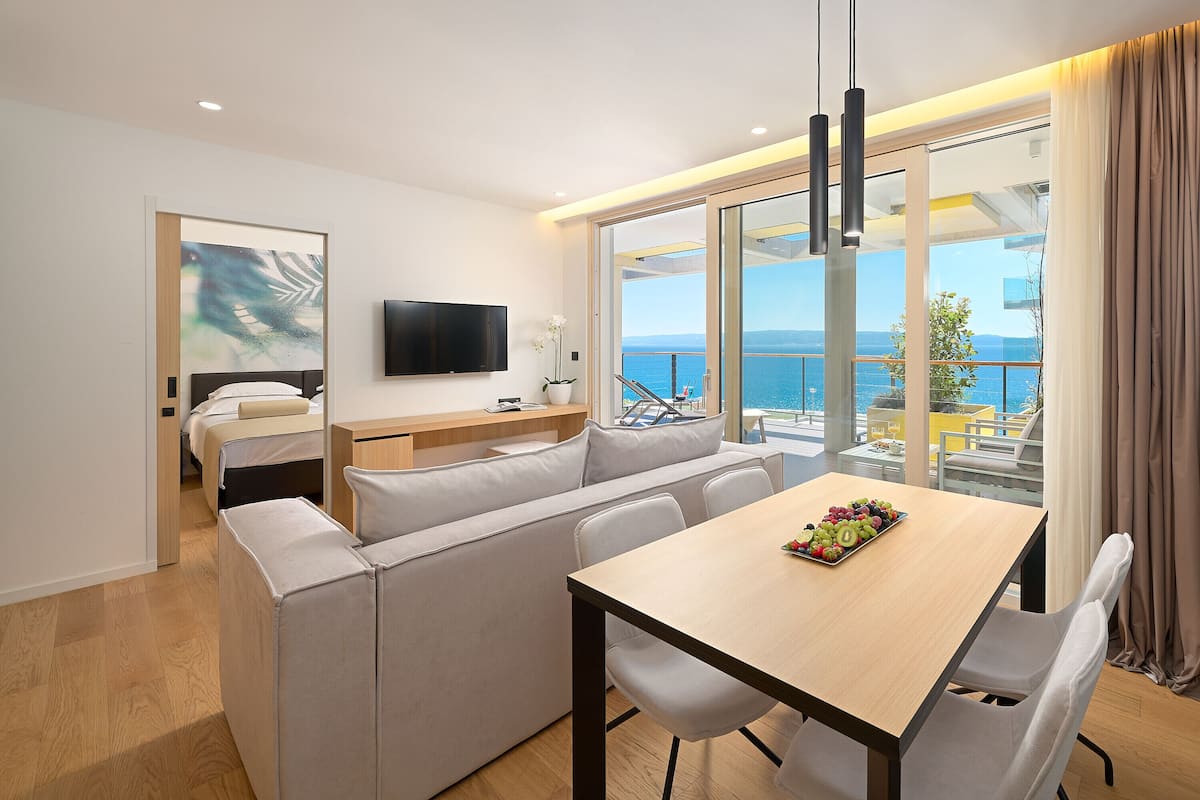Ein modernes Hotelzimmer mit einer Sitzecke, einem Esstisch und einem Fernseher. Es verfügt über einen Balkon mit Meerblick.